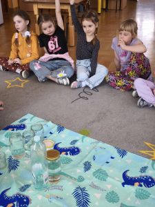 Dzieci siedzą na dywanie i porównują ilość wody w butelce i słoiku