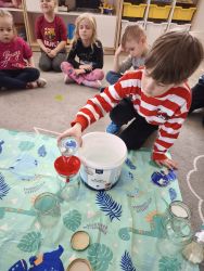 Chłopiec nabiera wodą do szklanki z wiaderka. Dzieci siedzą na dywanie