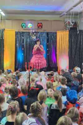Prowadząca Bal stoi na scenie ubrana w różową falbaniastą sukienkę, dzieci siedzą na dywanie.