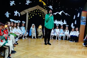 W tle dekoracja świąteczna- na ciemnym materiale stajenka Betlejemska, aniołki i gwiazdy. Dzieci ubrane w stroje pasterzy, aniołków, Maryi, Józefa, Królów i w stroje ludowe. Dzieci siedzą na krzesełkach a na środku stoi Pani dyrektor i mówi do mikrofonu.