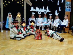 W tle dekoracja świąteczna- na ciemnym materiale stajenka Betlejemska, aniołki i gwiazdy. Dzieci ubrane w stroje pasterzy, aniołków, Maryi, Józefa, Królów i w stroje ludowe. Na środku sceny stoi sztuczne ognisko, dookoła niego siedzi 4 chłopców w strojach pasterzy
