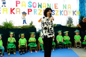 Dzieci ubrane w zielone stroje siedzą na krzesełkach. Na głowach mają papierowe birety. Na dekoracji widać napis Pasowanie na żaka przedszkolaka. Pani dyrektor stoi na środku i mówi do mikrofonu