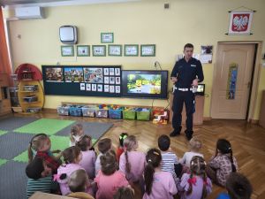 Dzieci siedzą na podłodze, przed nimi stoi policjant i pokazuje policyjną czapkę. W tle włączony telewizor z filmikiem ze spongeBob